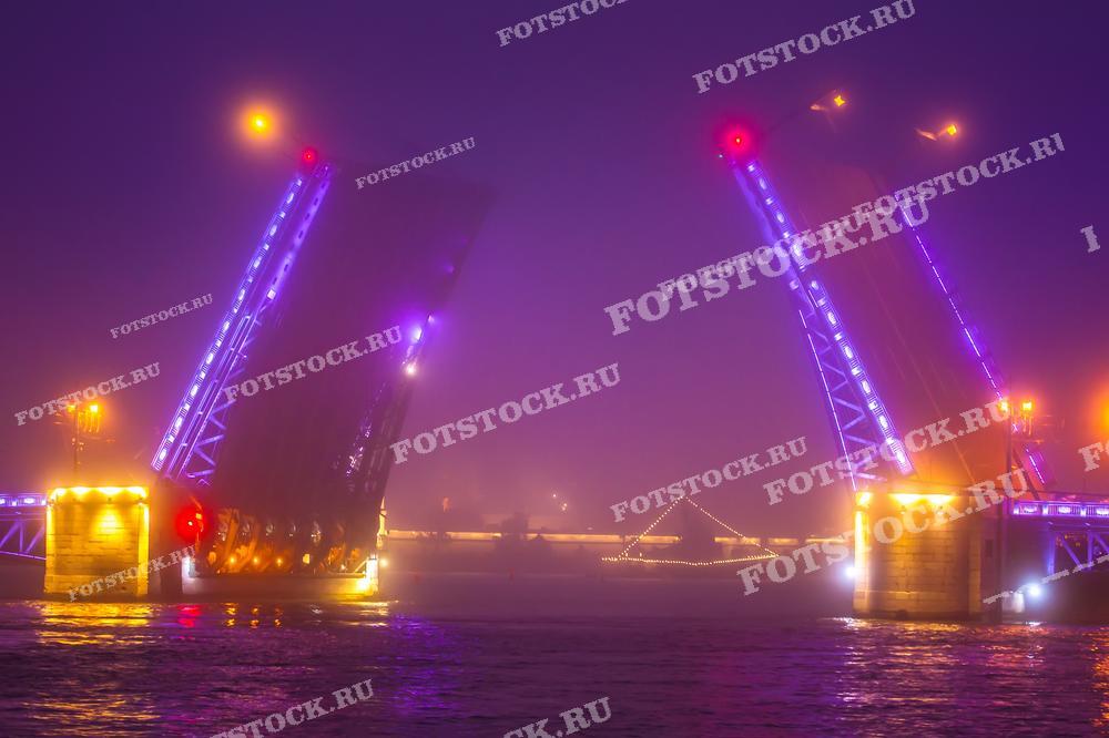 На фото изображен разводной мост ночью в Санкт-Петербурге. Видны яркие огни и отражения в воде, создающие атмосферу таинственности. Это фото подойдет для использования в туристических материалах, рекламных буклетах и статьях о Санкт-Петербурге.